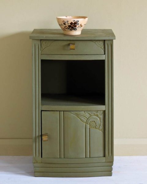 An olive green painted side table with carvings in the door and drawer. There is a small bowl on top.