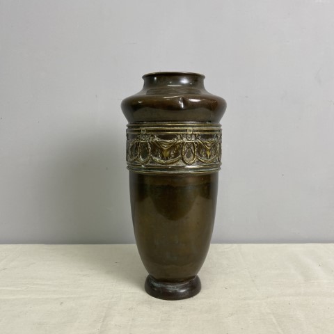 A tall brass vase with a wide opening at the top. It has raised decoration underneath the shoulder of the piece