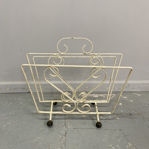 A white rectangular shaped wrought iron magazine rack with feet. It has decorative twirls and a carry handle