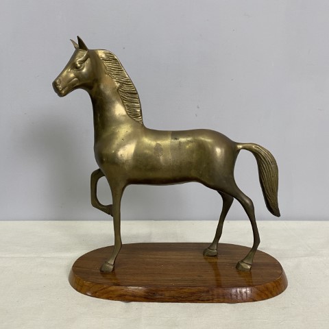 A brass statue of a standing horse with its front right leg raised. It is mounted on a timber base.