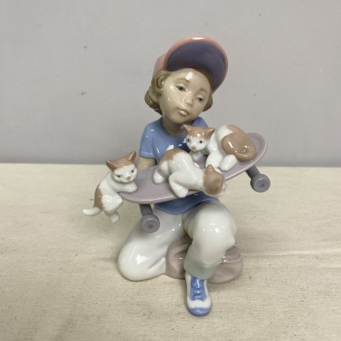 A Lladro figurine in pastel tones depicting a boy holding a skateboard with 3 kittens on it